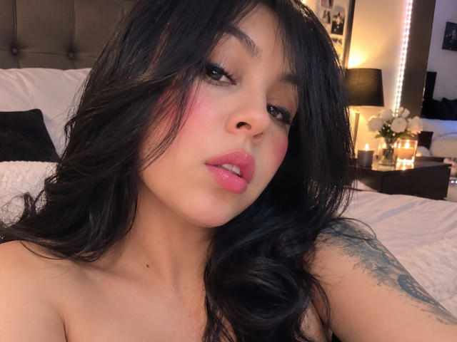NenaBlack heeft zwart, golvend haar, roze lippen en een neusring. Ze kijkt in de camera met lichtjes open roze lippen. Haar getatoeëerde arm is zichtbaar..