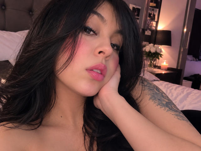 NenaBlack, com cabelo preto, lábios rosados e uma tatuagem no braço direito, apoiando o queixo na mão, olhando sedutoramente para a câmera em um quarto com pouca luz..