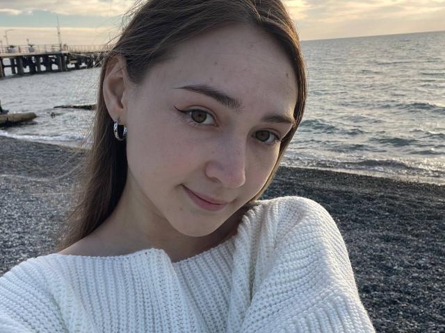 PearleSchoebel has light skin, green eyes, and brown hair, wearing a white knit sweater, with a small nose and a subtle smile, standing by a rocky beach at sunset.