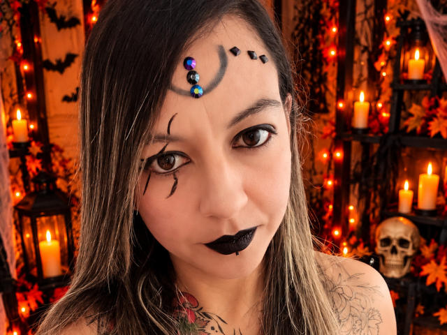 ArianaBlonde with black lipstick, dark eye makeup, and a face paint design with beads, has tattoos on her neck and shoulder, brown hair, and is in a Halloween-themed setting.