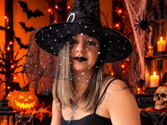 ArianaBlonde in a witch hat with star-patterned veil, black lipstick, and shoulder tattoos, smiling with medium tits and a black strap top, surrounded by Halloween candles and decor.