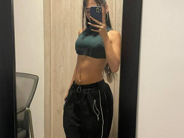 VioletaVelvet in a green sports bra, showing her abs and small tits, wearing black pants with white stripes, taking a mirror selfie with hands in pockets.