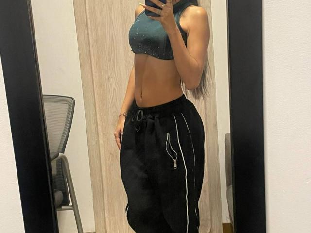 VioletaVelvet with a toned, tan, sexy, curvy body in a teal sports bra, showing her small tits and black, loose, zipped-up pants, taking a mirror selfie.