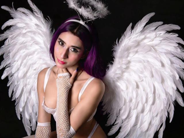 JuliaJobs, com cabelo roxo, asas de anjo brancas e um halo, vestindo luvas de rede de peixe e lingerie branca, tem a mão no queixo, olhando sedutoramente para a câmera..