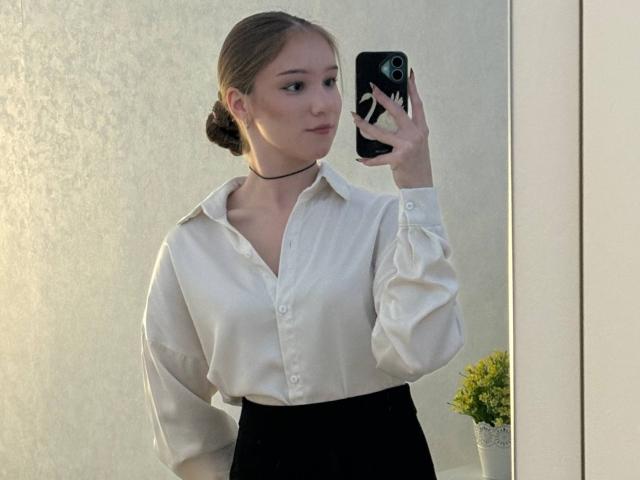 DomingaSierra con una camicia bianca e ampia, una gonna nera, una collana e i capelli intrecciati, che si scatta un selfie allo specchio, tenendo un telefono nero, mostrando un'espressione accigliata e una piccola pianta gialla..
