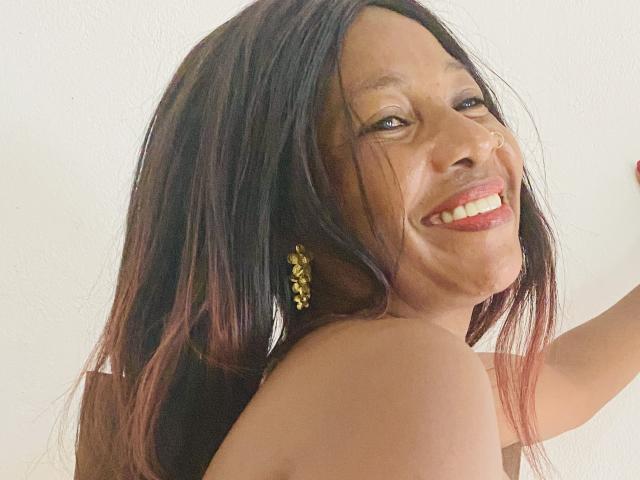 Hasina, con cabello largo de color marrón oscuro y puntas rojas, sonriendo con lápiz labial rojo, usando aretes amarillos, mostrando los hombros descubiertos, mirando por encima del hombro, sonriendo a la cámara..
