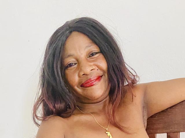 Hasina: Uma mulher com pele marrom escura, cabelo preto na altura dos ombros com pontas roxas, batom vermelho, sorrindo, colar de ouro, sem blusa, com o braço apoiado em uma cadeira de madeira, olhando para a câmera..