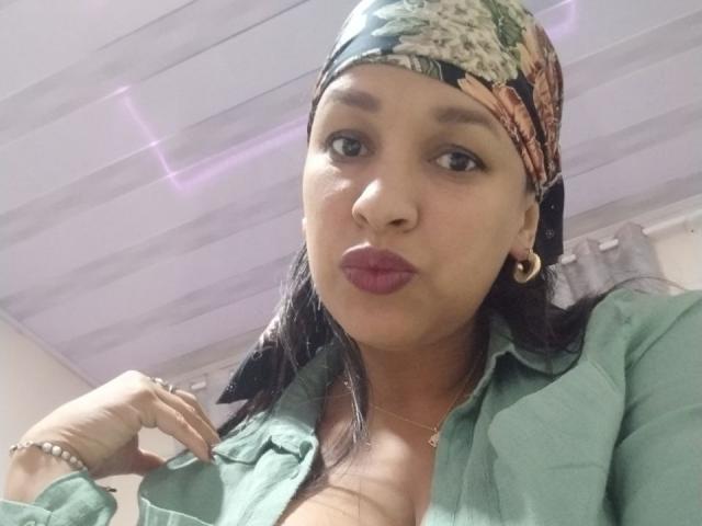 ZoehFerraro with brown skin, wearing a floral headscarf, green shirt, and gold hoop earrings, pouting with pink lipstick, showing cleavage, black hair, and a bracelet on her right hand.