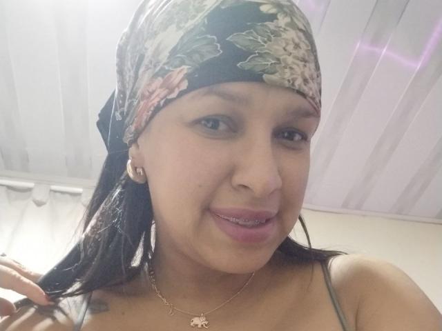 ZoehFerraro with floral headscarf, brown eyes, light brown skin, wearing a gold necklace with a small pendant, smiling, showing a slight overbite, touching her hair.