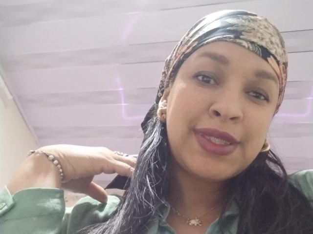 ZoehFerraro has a cute smile, dark eyes, and long black hair, wearing a patterned headscarf, green shirt, and bracelet, with purple lipstick and a necklace.