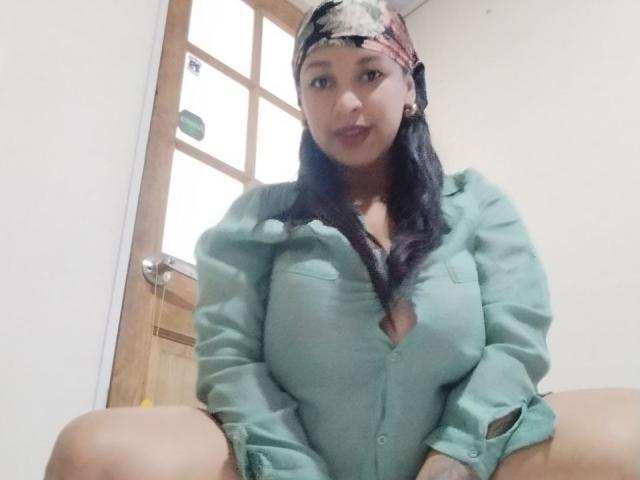 ZoehFerraro with dark hair, wearing a green shirt, patterned headscarf, and dark lipstick, sitting with legs spread, showing her tits and pussy slightly. Wooden door in background.