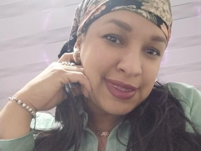 ZoehFerraro has medium brown skin, long dark hair, and is wearing a floral headscarf, green shirt, and pink lipstick, with a bracelet and necklace, smiling at the camera.