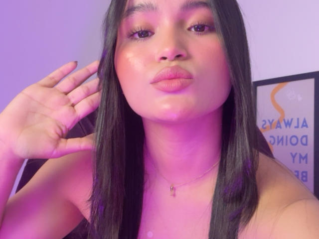 MiiaPalmer with long black hair, full pink lips, shiny skin, wearing a necklace, making a kissing face, hand near her head, in purple and pink lighting.