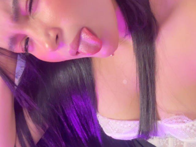 MiiaPalmer with long black hair, sticking out her shiny pink tongue, wearing a white lace bra, purple light on her face, and a small bead on her neck.