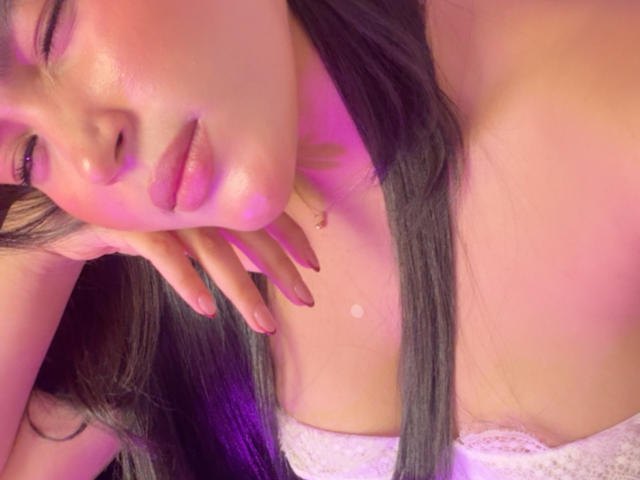 MiiaPalmer met glanzende, roze lippen, lang donker haar en roze-paarse verlichting, leunt haar wang op haar hand en laat haar witte, met kant bedekte borsten zien. Haar nagels zijn rood..