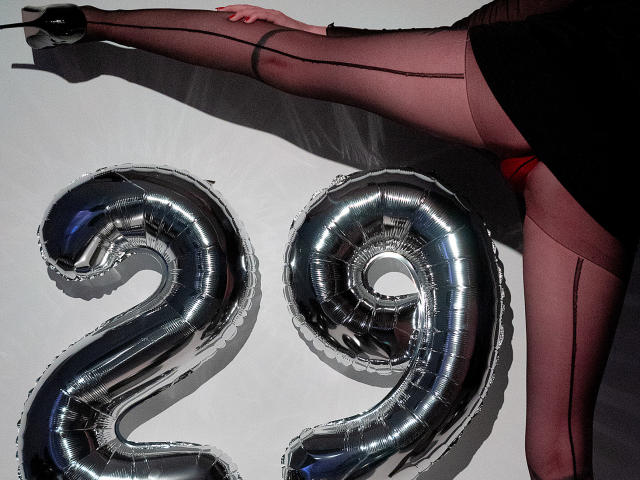 La gamba di AlesandraLovi, con tacchi alti neri e collant rosa trasparenti, è piegata sopra dei palloncini metallici lucidi a forma di "29". Il suo sedere è parzialmente visibile, indossa uno slip rosso..