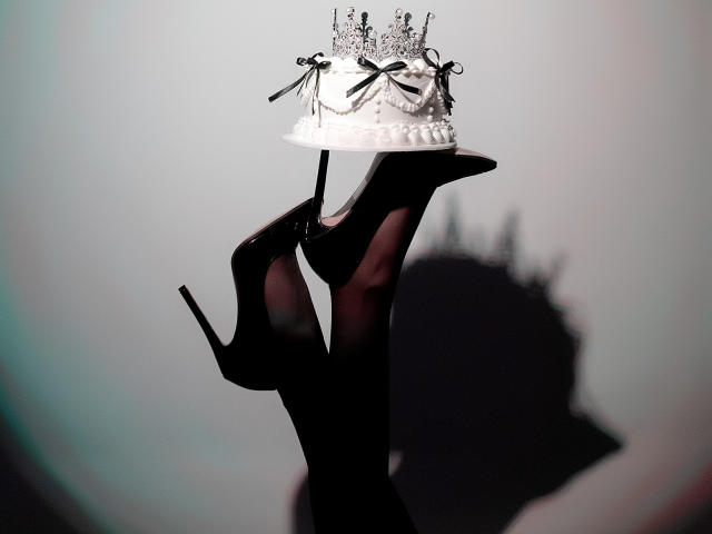 AlesandraLovis schwarze Stilettos halten eine weiße, mit Diamanten besetzte Torte mit schwarzen Bändern vor einem grauen Hintergrund, wobei ihre Absätze einen Schatten werfen..
