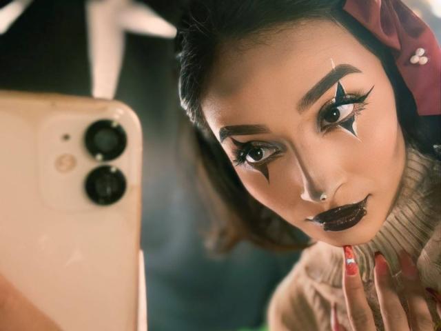 LunaixMoon's face with star-shaped eye makeup, dark lipstick, and red hair bow, taking a selfie with a gold iPhone, showing off manicured red nails.