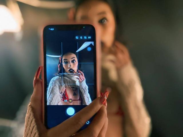 LunaixMoon with red nails and a sweater, holding a phone showing a selfie of a woman with large tits, wearing headphones and a white sweater, in a dim room.