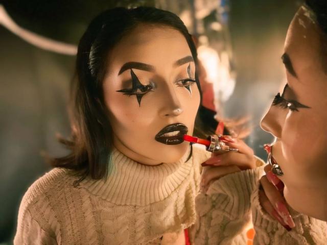 LunaixMoon has dark makeup with star-shaped eyeliner, black lipstick, and a red lipstick applicator in her mouth, wearing a beige knitted turtleneck, reflected in a mirror.