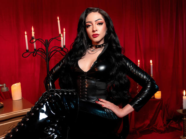 Alesandra Lovi, con lunghi capelli neri, indossa un abito e un corsetto di lattice nero lucido, mostrando il seno, seduta in una stanza con candele e tende rosse..