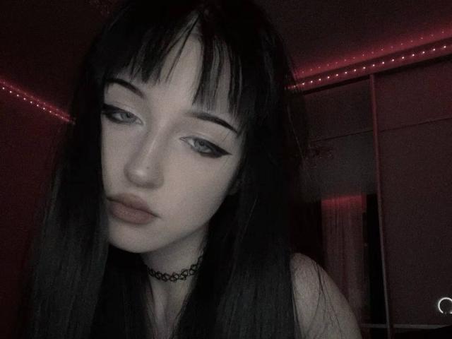 KiraLowkey with pale skin, dark straight hair, black eyeliner, and a choker, looking down with pouty lips. Red LED strip lights in the background.