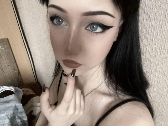 KiraLowkey with pale skin, black eyeliner, blue eyes, and black nails, touching her chin with long black nails, wearing a black strap, looking at the camera.