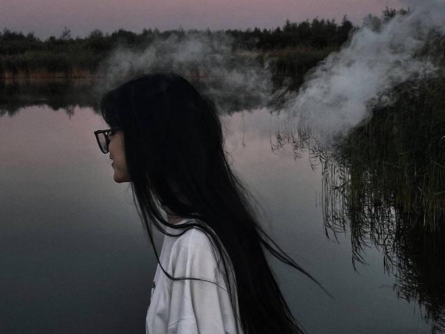 KiraLowkey with long black hair, glasses, white shirt, and steam rising from her mouth, standing by a calm, reflective water body at dusk.