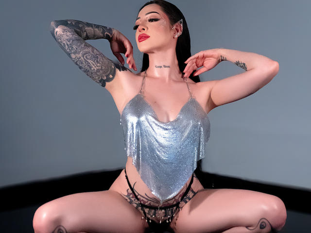 AlesandraLovi with pale skin, black tattoos, and red lipstick, wearing a silver, shimmering, barely-there top, and black fringed panties, squatting with arms raised, showing off her tits.