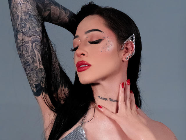 Alesandra Lovi com tatuagens, batom vermelho, brilho nas bochechas, presilha de cabelo prateada, unhas vermelhas, mão no pescoço, braço levantado, mostrando tatuagens no braço, fundo cinza e blusa prateada brilhante..