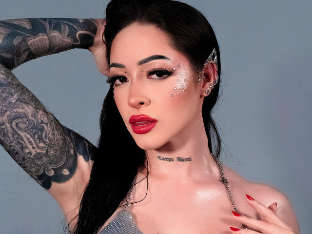 AlesandraLovi tiene tatuajes, lápiz labial rojo y brillantina en la mejilla. Está levantando el brazo, mostrando su tatuaje en la manga. Lleva un collar de cadena y tiene las uñas rojas..