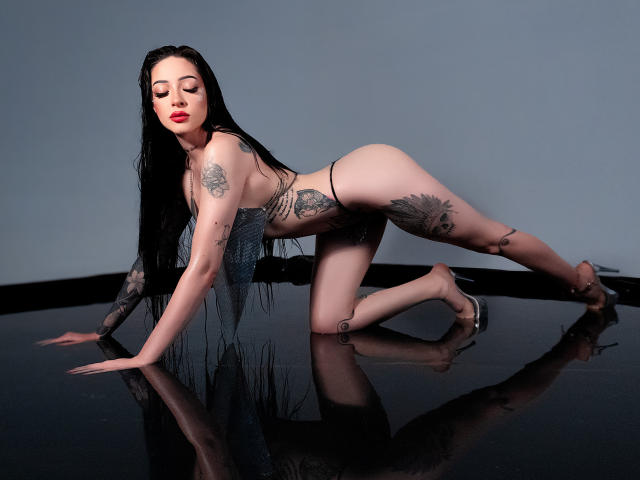AlesandraLovi on all fours, wearing a sheer black top, revealing her tits and ass. She has tattoos on her arm, side, and thigh, with long black hair.