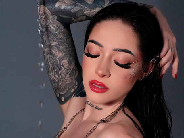 AlesandraLovi, avec ses cheveux foncés, son rouge à lèvres rouge, son bras tatoué et son maquillage scintillant, les yeux fermés, le bras levé, mouillée, et portant un haut en chaîne, avec des gouttes d'eau..