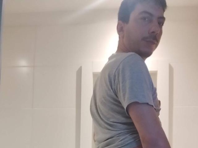XSantyX, con cabello y bigote oscuros y cortos, vistiendo una camiseta gris, mirando por encima del hombro, en una habitación ligeramente tenue. Su trasero es parcialmente visible..