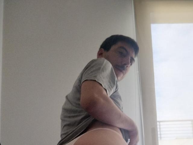 XSantyX with short dark hair, gray t-shirt, and white underwear, looking over shoulder, exposing pale, round ass. Soft lighting, slightly blurred. Slightly chubby, casual pose.