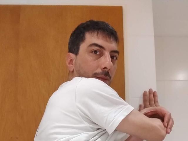 XSantyX, cheveux courts et foncés, moustache, t-shirt blanc, regardant par-dessus son épaule, bras croisés, porte en bois derrière lui, expression sérieuse, teint de peau moyen, légèrement musclé, mur blanc à droite..