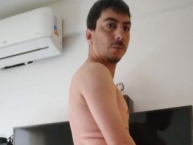 XSantyX senza camicia, con un baffo, capelli scuri e corti e un leggero sorriso, che guarda oltre la spalla. Il suo petto e le sue braccia sono visibili, con un condizionatore d'aria sopra..