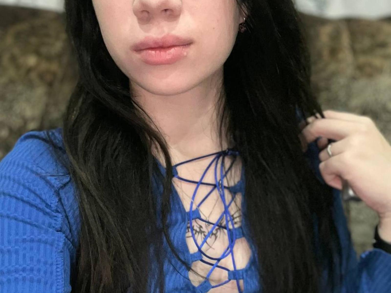 Il viso di Kenneth Verma, con la pelle pallida, le labbra rosate e i lunghi capelli neri, indossa una maglia blu a maglia con un design allacciato che mette in mostra il décolleté. Una mano è vicina al viso, con un anello..