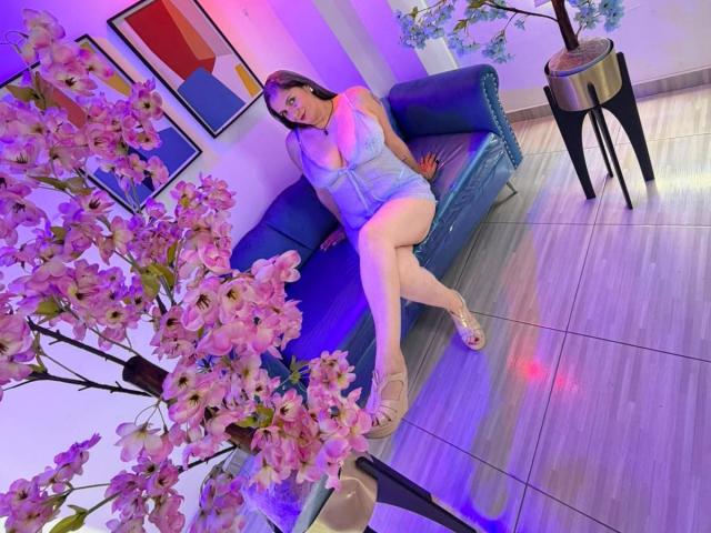 Madame Scarlett, con un seno prosperoso, indossa un abito blu e tacchi beige, seduta su un divano blu con fiori di ciliegio rosa e luci viola sullo sfondo..