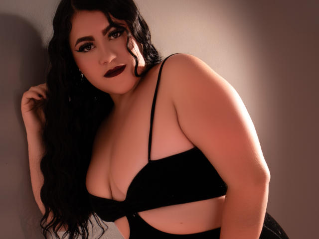 ArieWilden, con capelli scuri e ondulati, rossetto nero e reggiseno nero, che mostra un seno generoso, in piedi, guarda la fotocamera, con una mano tra i capelli, luce soffusa, sexy, sicura di sé..