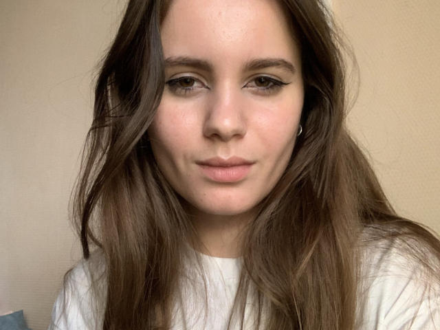 Nella Koczur, avec ses longs cheveux bruns, sa peau claire et son maquillage discret, porte une chemise blanche, a une expression boudeuse et un nez légèrement retroussé, et regarde directement l'appareil photo..