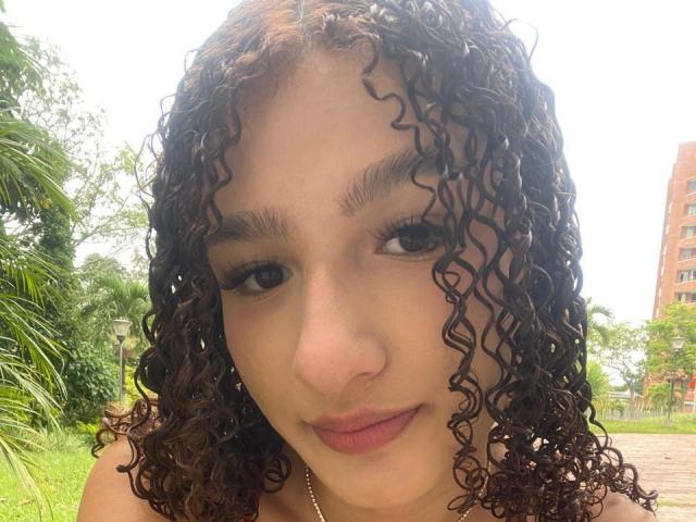 Nathacha Evans ha i capelli castano ricci, un naso marrone chiaro e labbra leggermente divisa con un sorriso appena accennato. Sta guardando di lato con un'espressione calma..