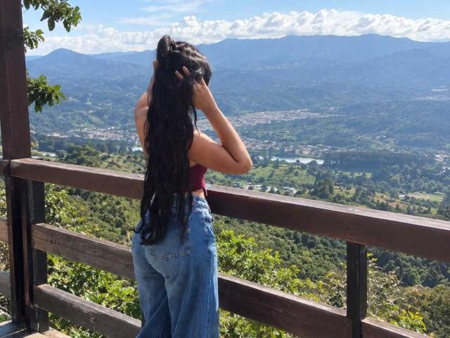 AmyLambert with long black hair, wearing a red top and blue high-waisted jeans, stands by a wooden fence, looking at a mountainous landscape, her ass slightly visible.