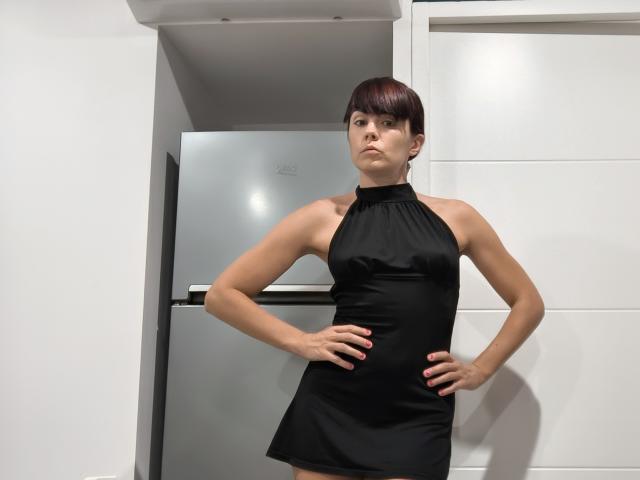 OrianaLaFrancaise steht in einem schwarzen Top mit Trägern, die Hände sind auf den Hüften, sie hat kurze, braune Haare, einen ernsten Gesichtsausdruck, ihre Arme sind entblößt, sie hat kleine Brüste, im Hintergrund ist eine weiße Wand und ein Kühlschrank zu sehen, es gibt kaum Schatten..