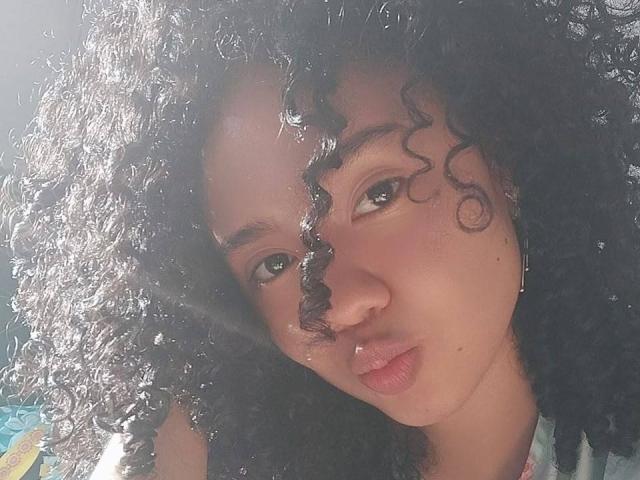 KirasPell with curly black hair, brown skin, and pouty lips, looking to the side. Sunlight shines through her hair, highlighting her face and small nose.