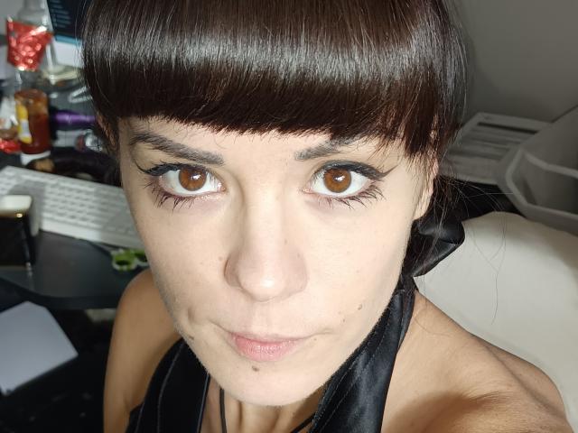 OrianaLaFrancaise hat braune Augen, kurze, dunkelbraune Haare mit Pony und trägt schwarzen Eyeliner. Sie schaut in die Kamera und lächelt leicht, wobei ihre Lippen geschlossen sind. Ihre Brüste sind nicht sichtbar..