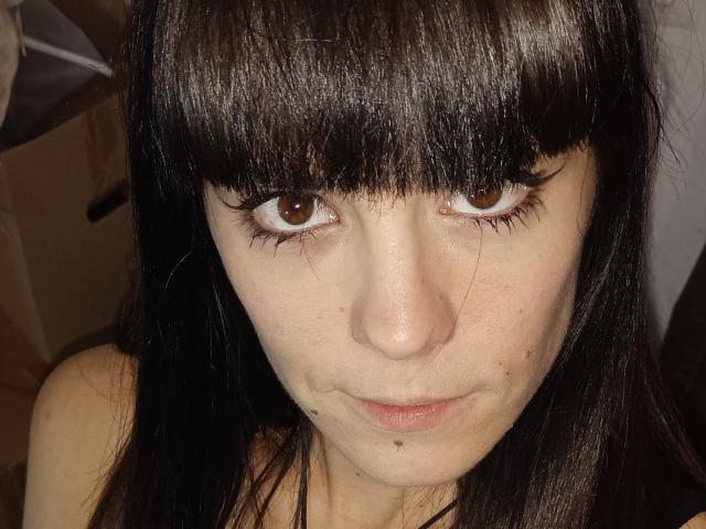 OrianaLaFrancaise, con occhi marrone scuro, frangia nera e capelli neri lisci, guarda la fotocamera con un'espressione leggermente seria. Ha la pelle chiara con piccole lentiggini..