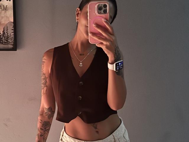 MajoJones con tatuaggi, indossa un gilet nero senza maniche, pantaloni bianchi, tiene in mano un telefono rosa, porta un orologio intelligente al polso, sta scattando un selfie allo specchio, mostrando un torso snello e tonico e un addome piatto..