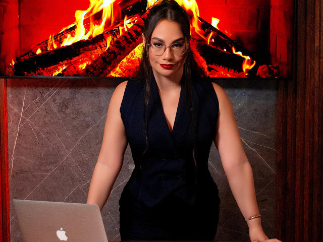 Emma Ricco, con rossetto rosso, occhiali e vestito nero, tiene in mano un portatile argentato e si trova di fronte a un camino rosso-arancio con legna che brucia. Ha una figura formosa e sensuale..