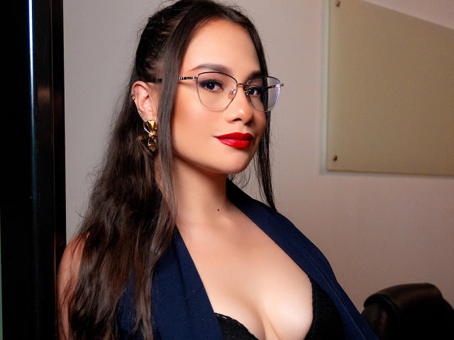 Emma Ricco, con gli occhiali, il rossetto rosso e il gilet blu scuro, mostra il seno in un reggiseno nero, indossa orecchini dorati, ha i capelli castani lunghi e un leggero sorriso..